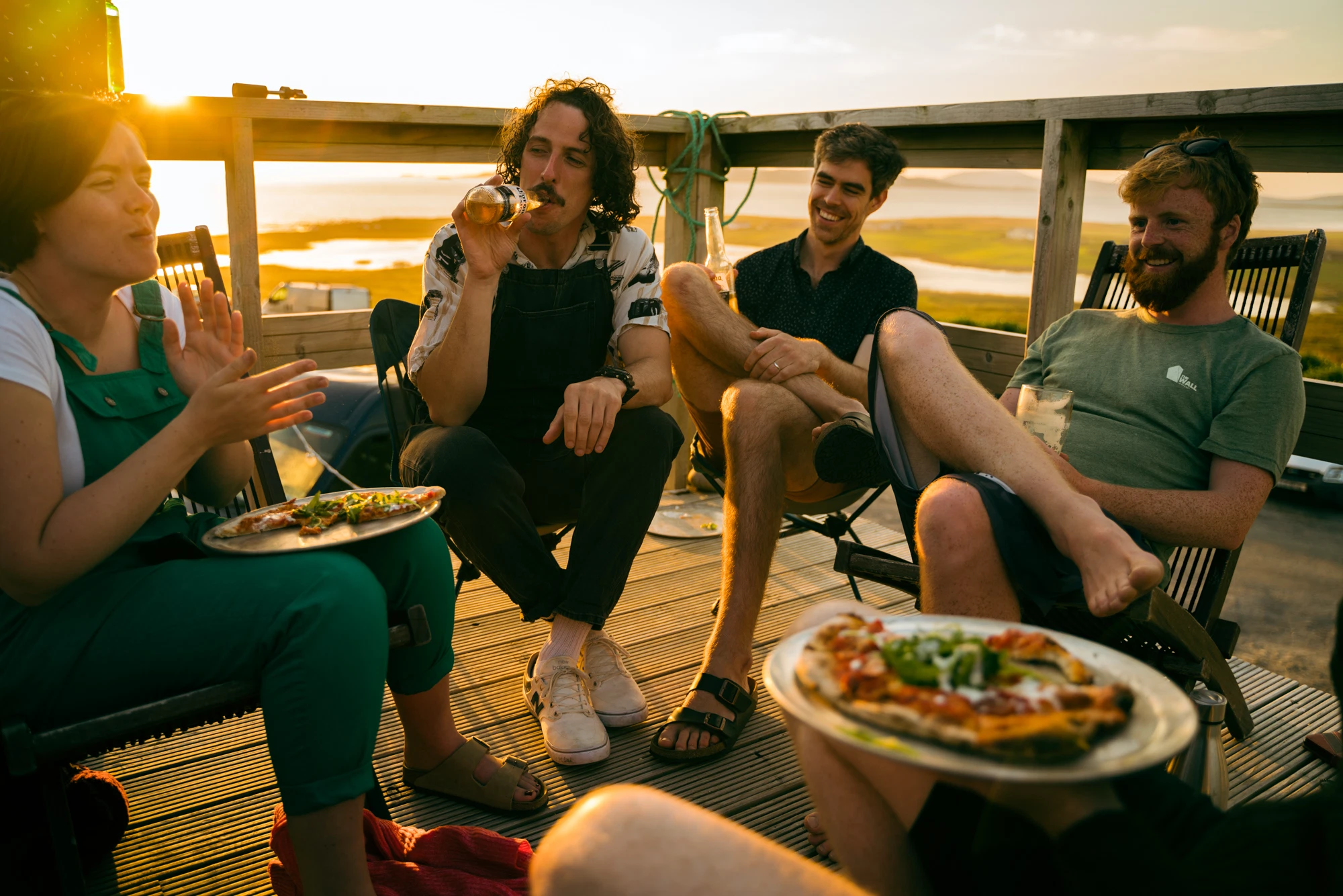 Friends at sunset, enjoying pizza and drinks on a wooden deck by the sea, creating a relaxed atmosphere.