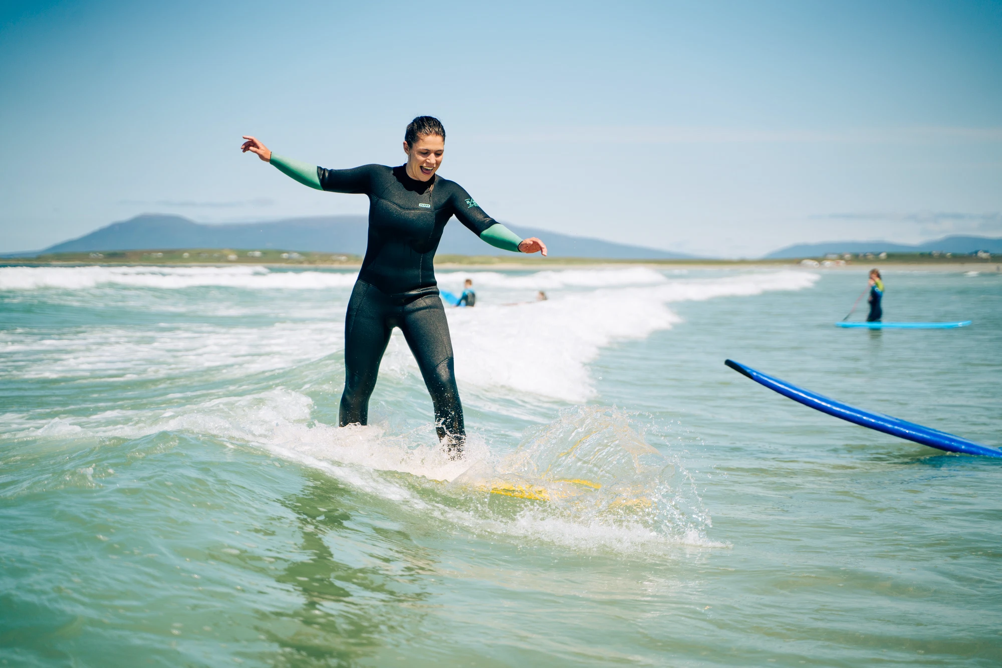 Woman surfing on a wave in the ocean, wearing a wetsuit, with distant mountains in the background.