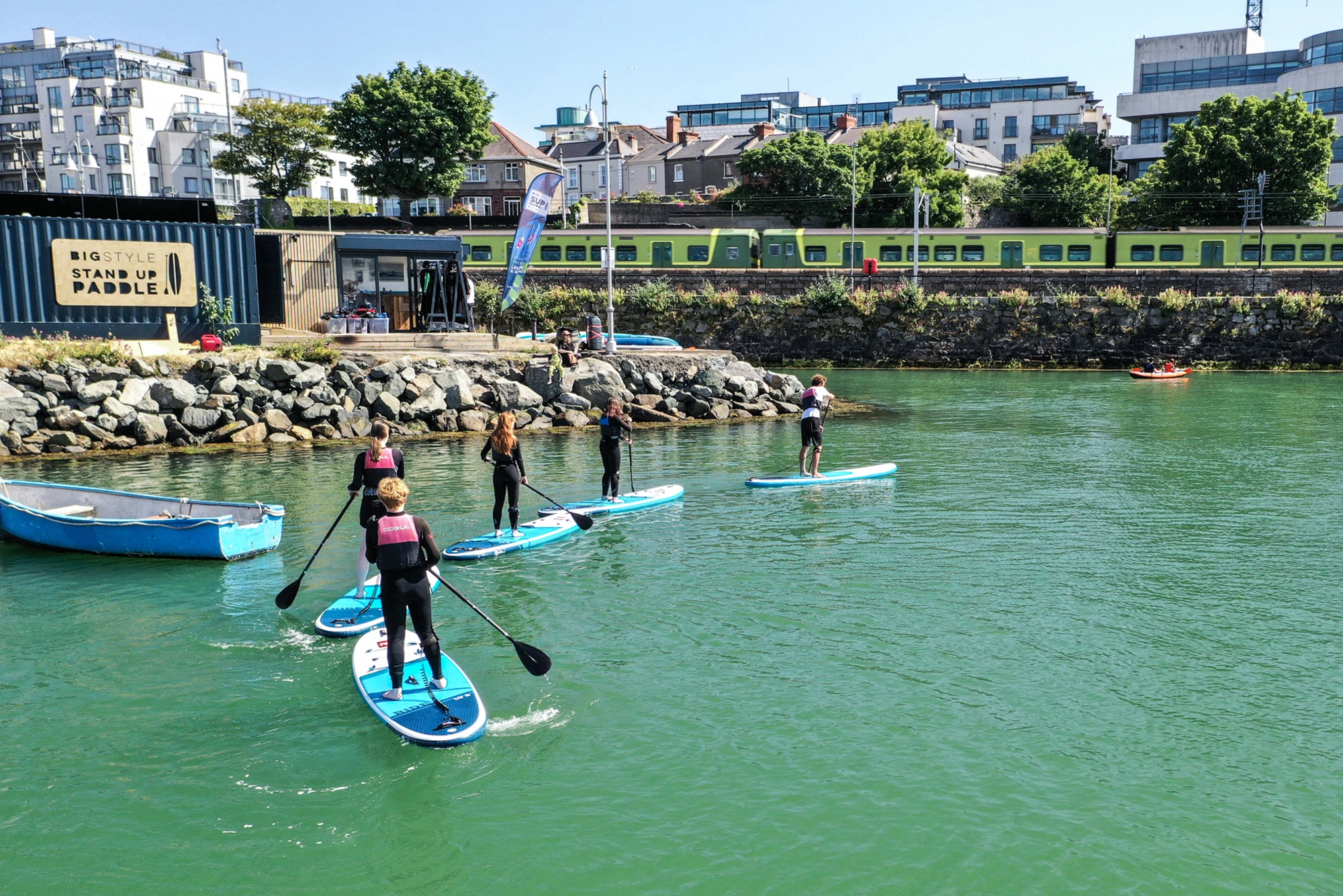 Group paddleboarding in Dublin bay, sunny day; train and buildings in background. Outdoor water sports.