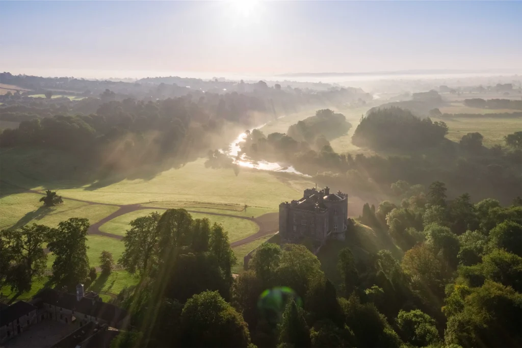 Aerial view of an Irish castle surrounded by misty fields and sunlight streaming through trees at dawn.