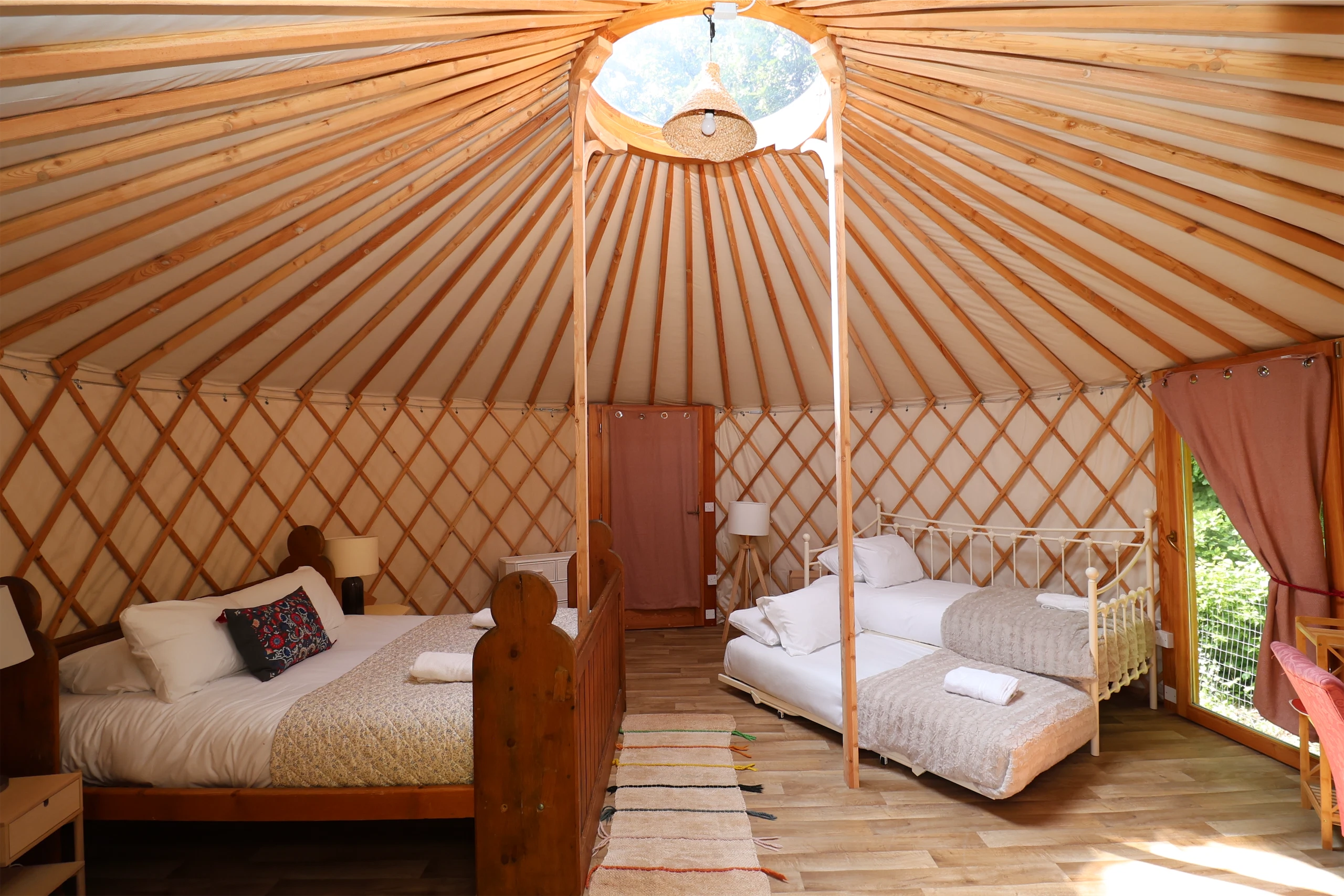 Cosy yurt interior with double and single beds, wooden floor, natural light, and a circular roof window.