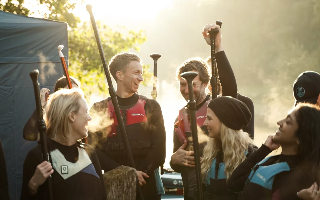 Group of friends in wetsuits with paddles, chatting and laughing outdoors in sunlight.