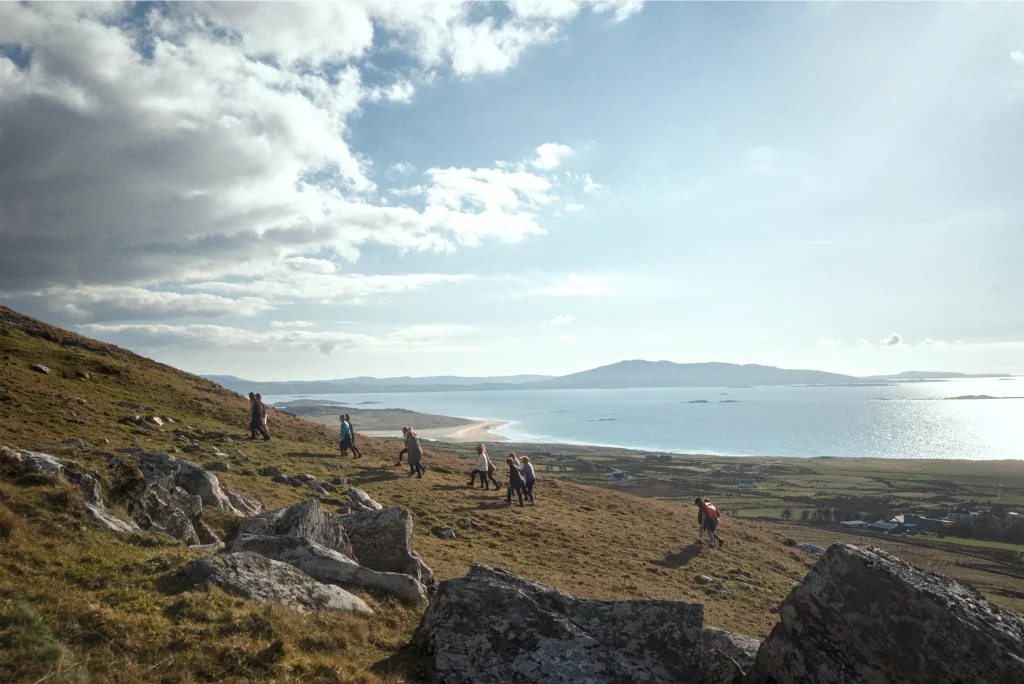 Hikers on a grassy hill overlooking the beach and sea under a cloudy sky in Donegal, Ireland.