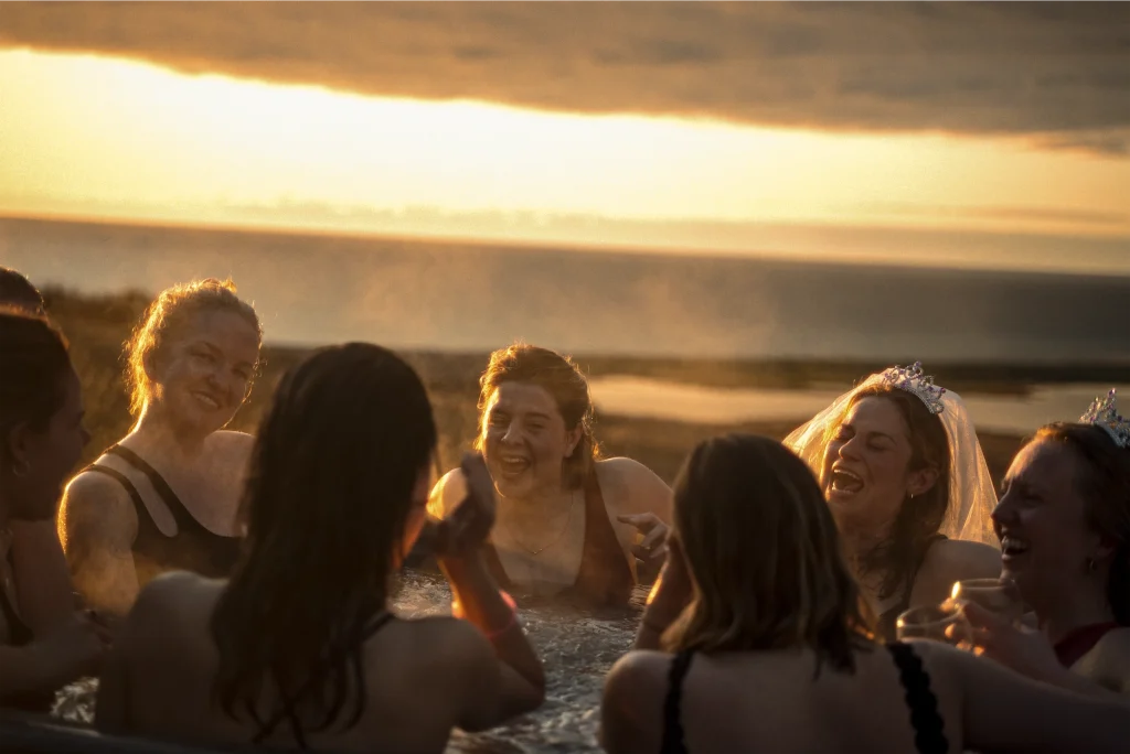 Women in swimsuits enjoy a hot tub at sunset, laughing and wearing tiaras, with a scenic water view.