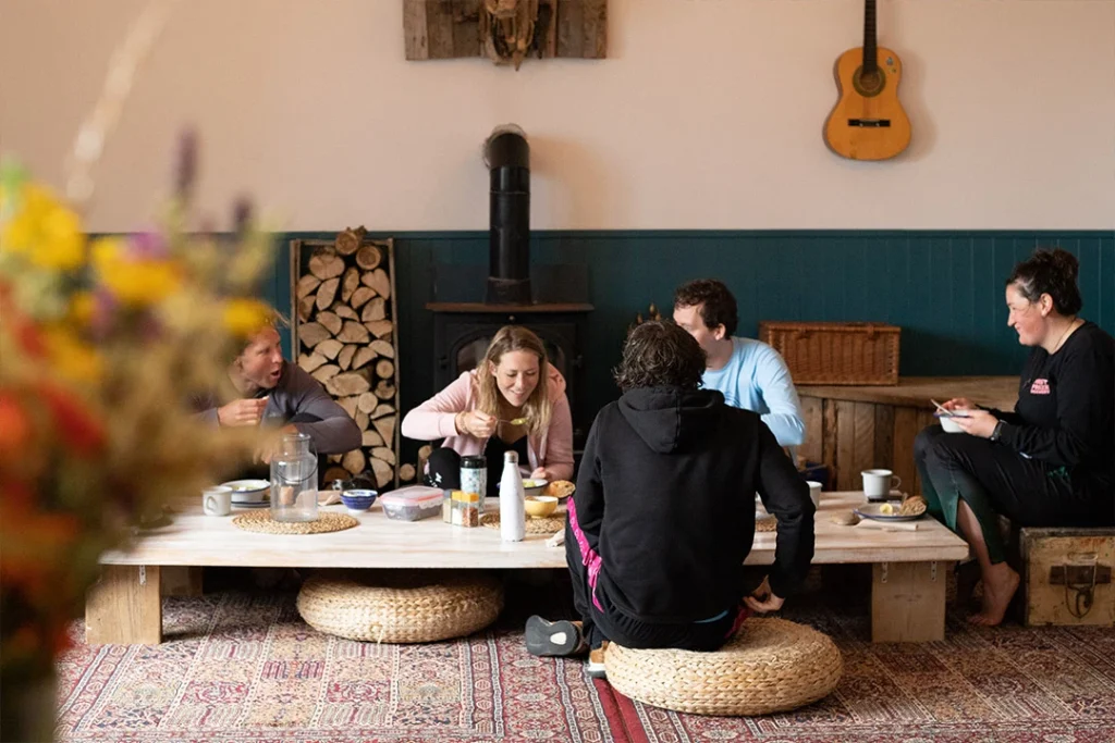 Group dining around low table with cushions in cosy room; wood burner and guitar decor.