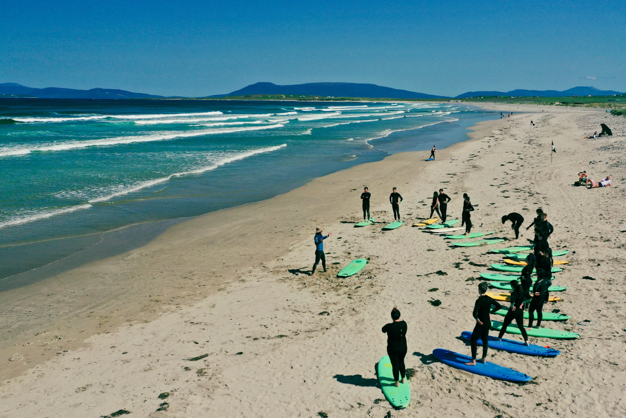 Surfers in wetsuits with boards on a sandy beach, waves in the background, clear sky, remote Irish coastline scene.