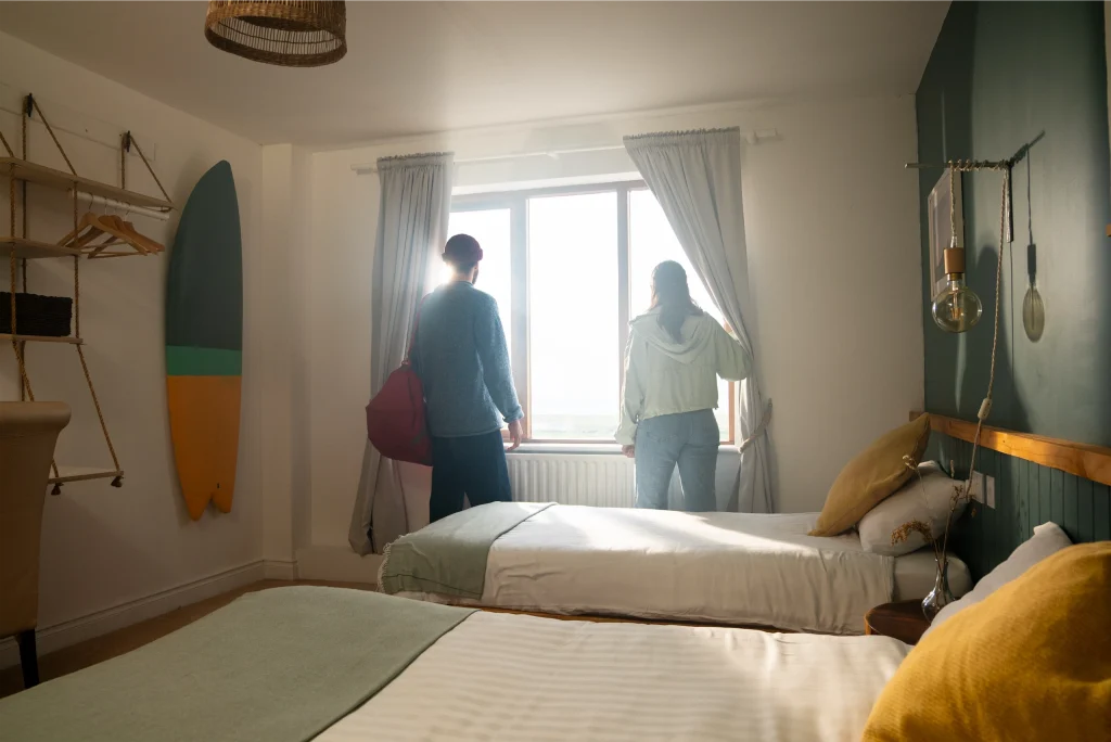 Two people look out a window in a cosy bedroom with twin beds and a surfboard on the wall.