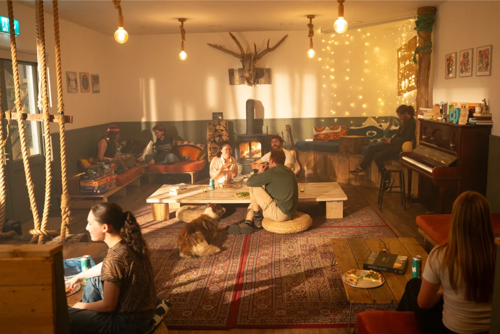 Cosy lounge with people chatting, sitting on cushions around a table. Warm lighting, piano, dog, and board games.