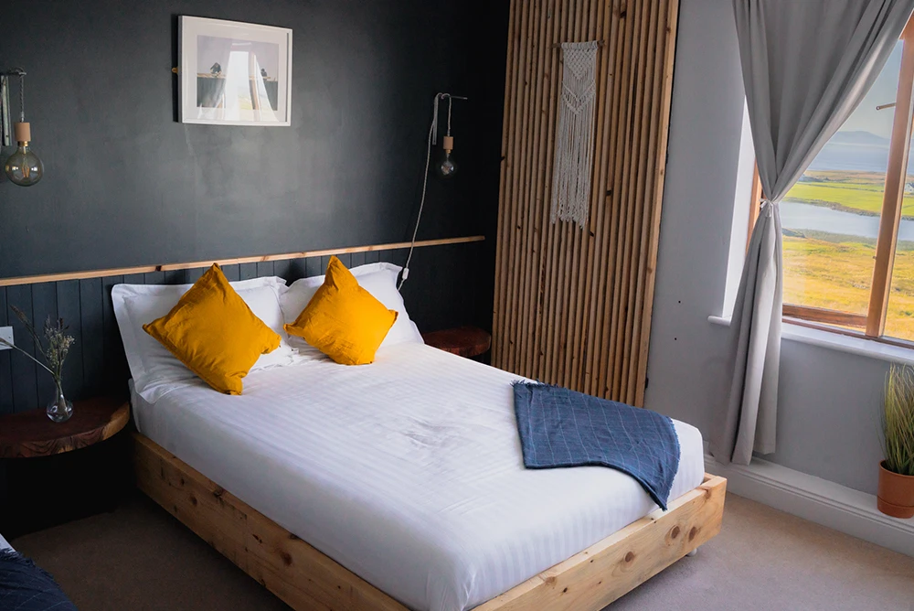 Cosy bedroom with wooden bed, yellow cushions, dark feature wall, and window view of scenic landscape.