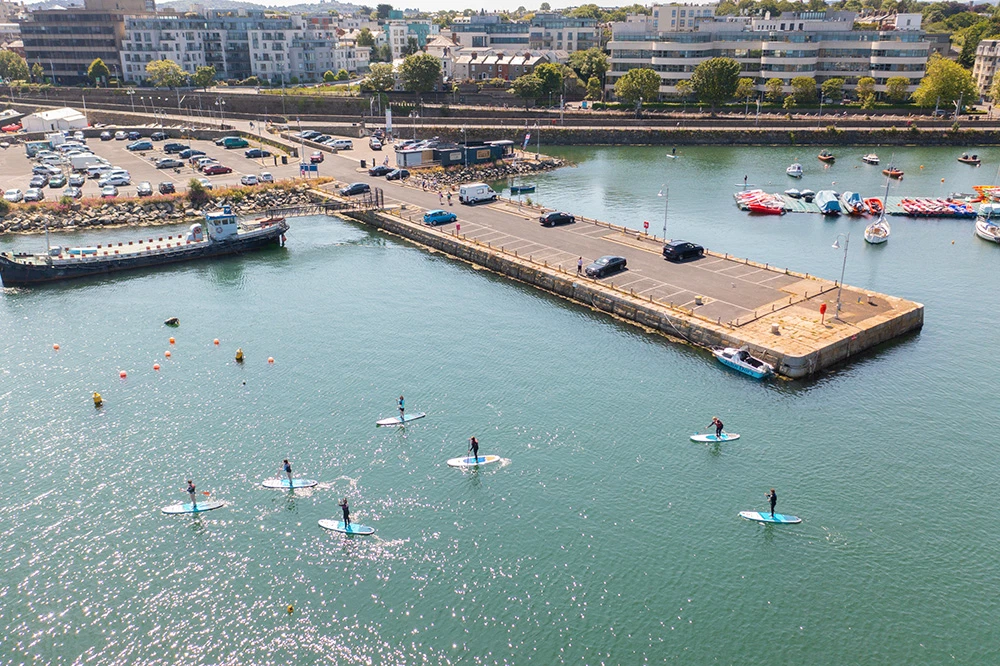 Aerial view of paddleboarders in a marina, surrounded by boats and waterfront buildings on a sunny day.