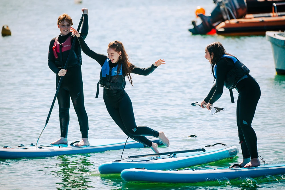 Three people in wetsuits paddleboarding, one playfully jumping, on a sunny day at sea.