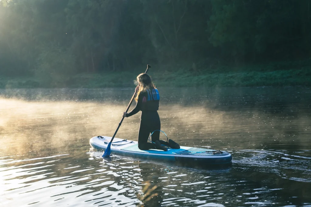 Woman kneeling on paddleboard at sunrise with misty forest backdrop; peaceful outdoor water activity scene.