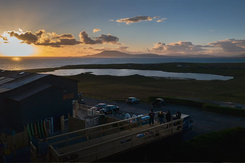 Sunset view over Achill Island, Ireland, with people on a deck, ocean and mountain in the background.