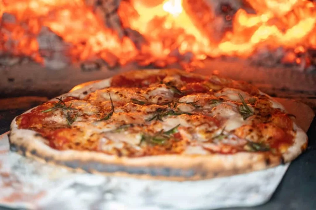 Wood-fired pizza with melted cheese and herbs baking in a traditional oven, flames glowing in the background.
