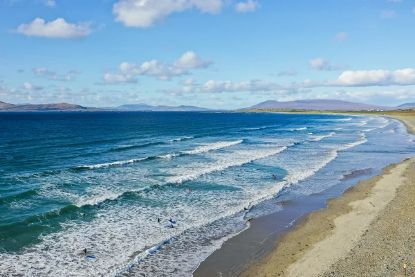 Irish beach with surfers, calm waves, blue sky, and distant mountains; perfect for seaside activities and views.