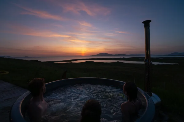 Three people relax in a seaside hot tub at sunset, overlooking scenic mountains and sea in County Mayo, Ireland.