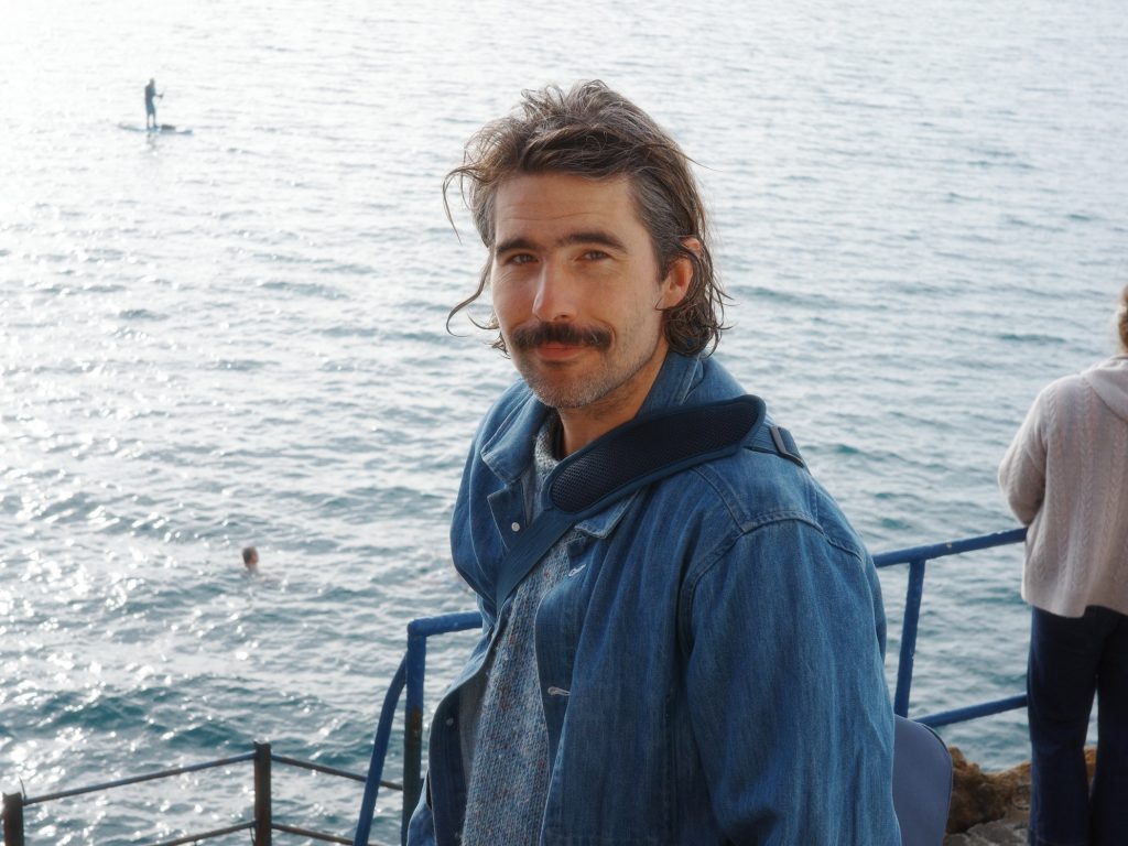 Man in denim jacket by the sea, with paddleboarder and swimmer in the background.