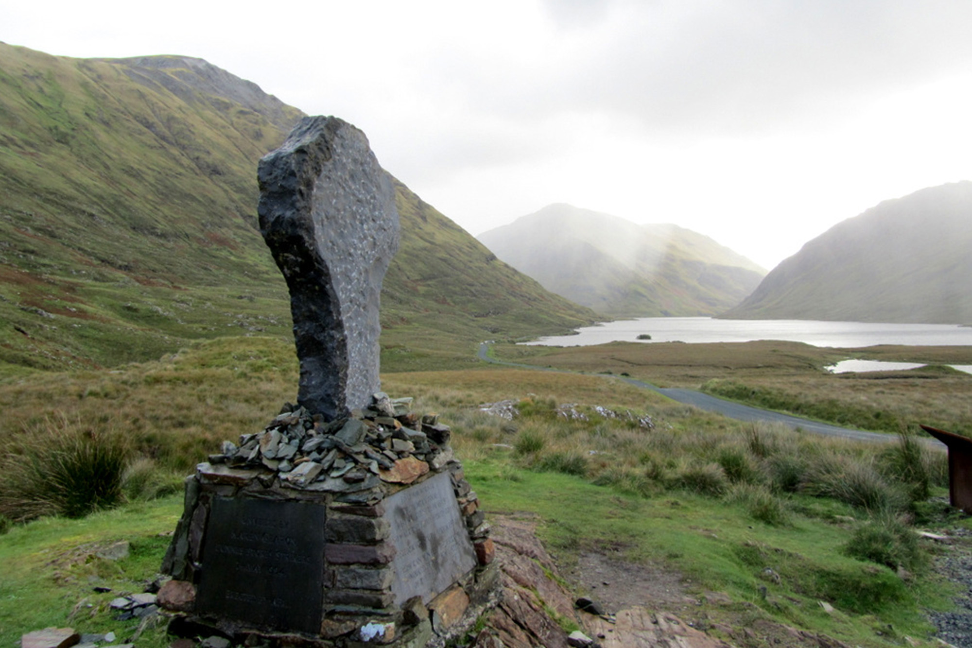 Stone memorial in scenic Irish valley with mountains and a lake under a cloudy sky.
