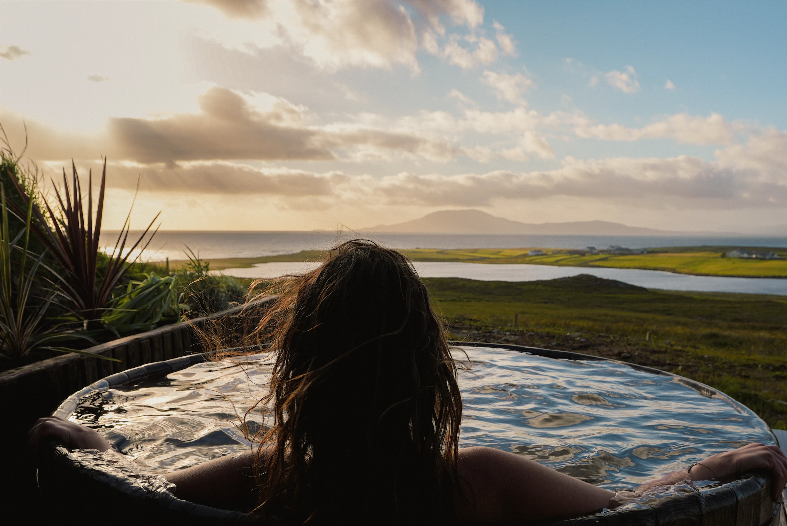 Woman relaxing in outdoor hot tub overlooking scenic Irish landscape at sunset.