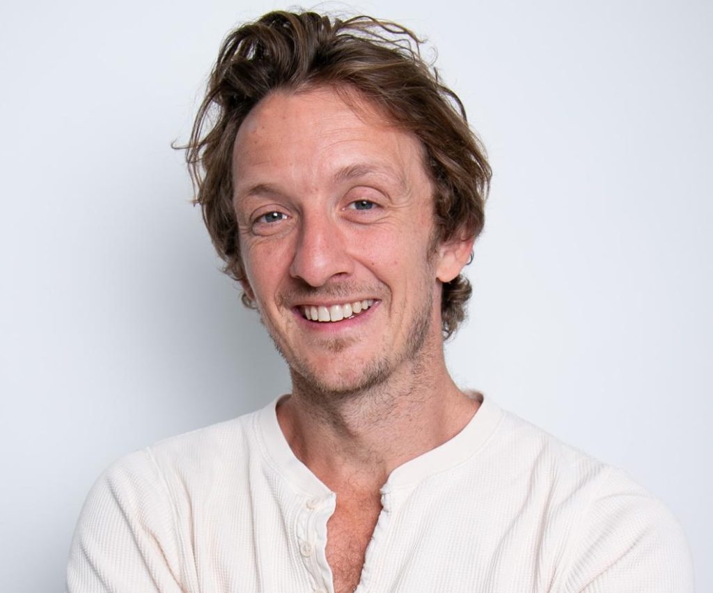 Smiling man in a white shirt with a relaxed hairstyle on a plain background.