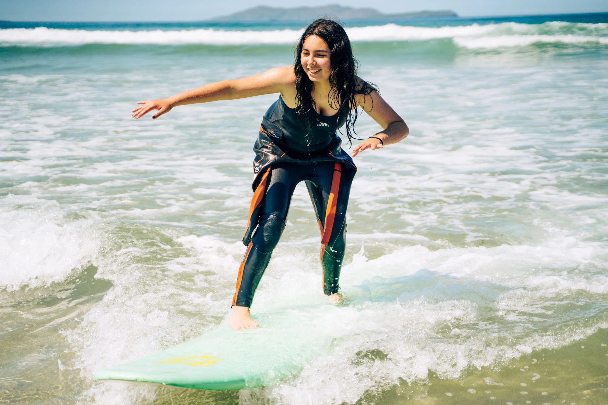 Young woman surfing on a sunny day, balancing on a green surfboard with waves in the background.
