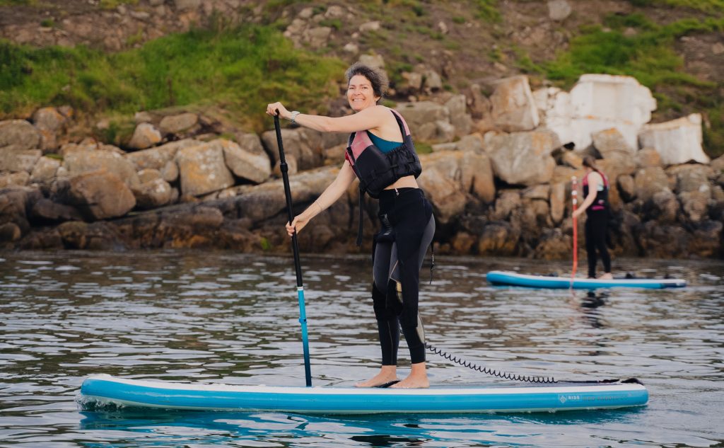 Woman paddleboarding on calm sea near rocky shoreline, wearing wetsuit and buoyancy aid.