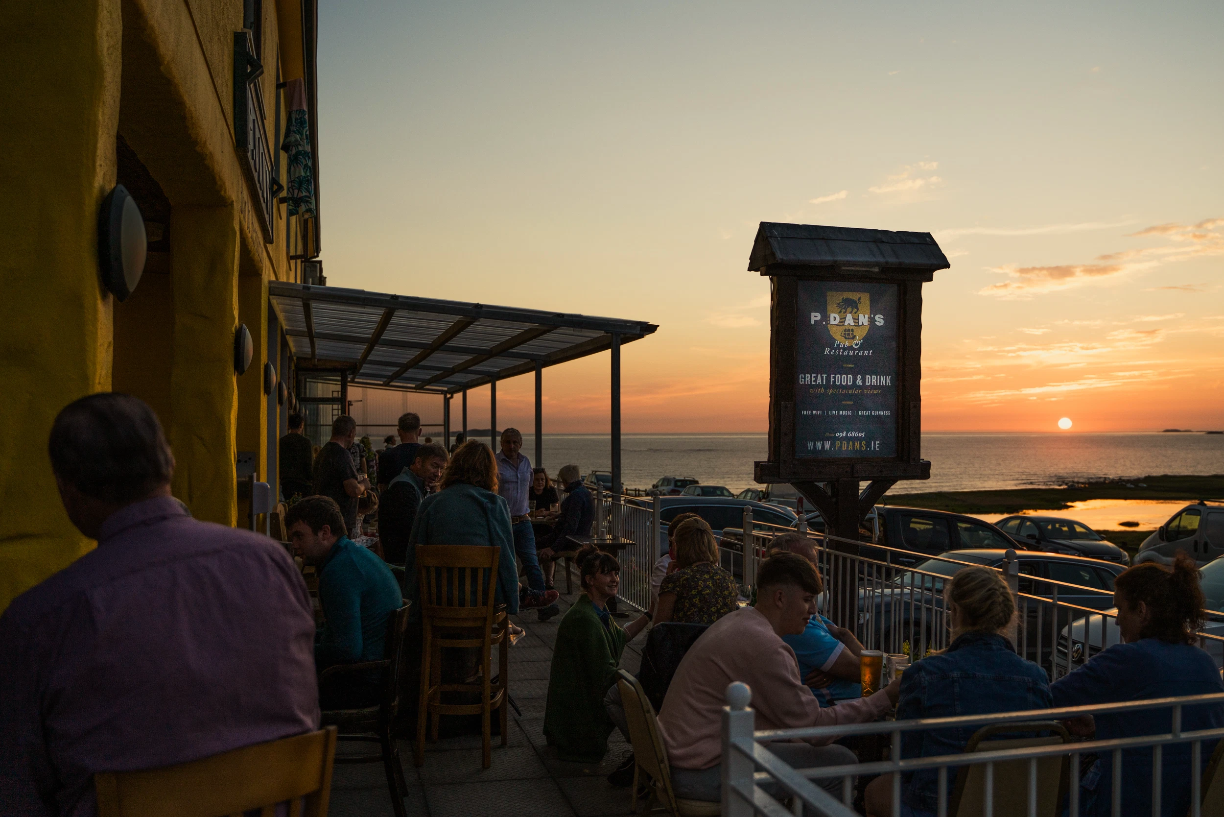 Pub patrons seated outdoors at sunset, overlooking the sea, with a sign for P. Daan's.