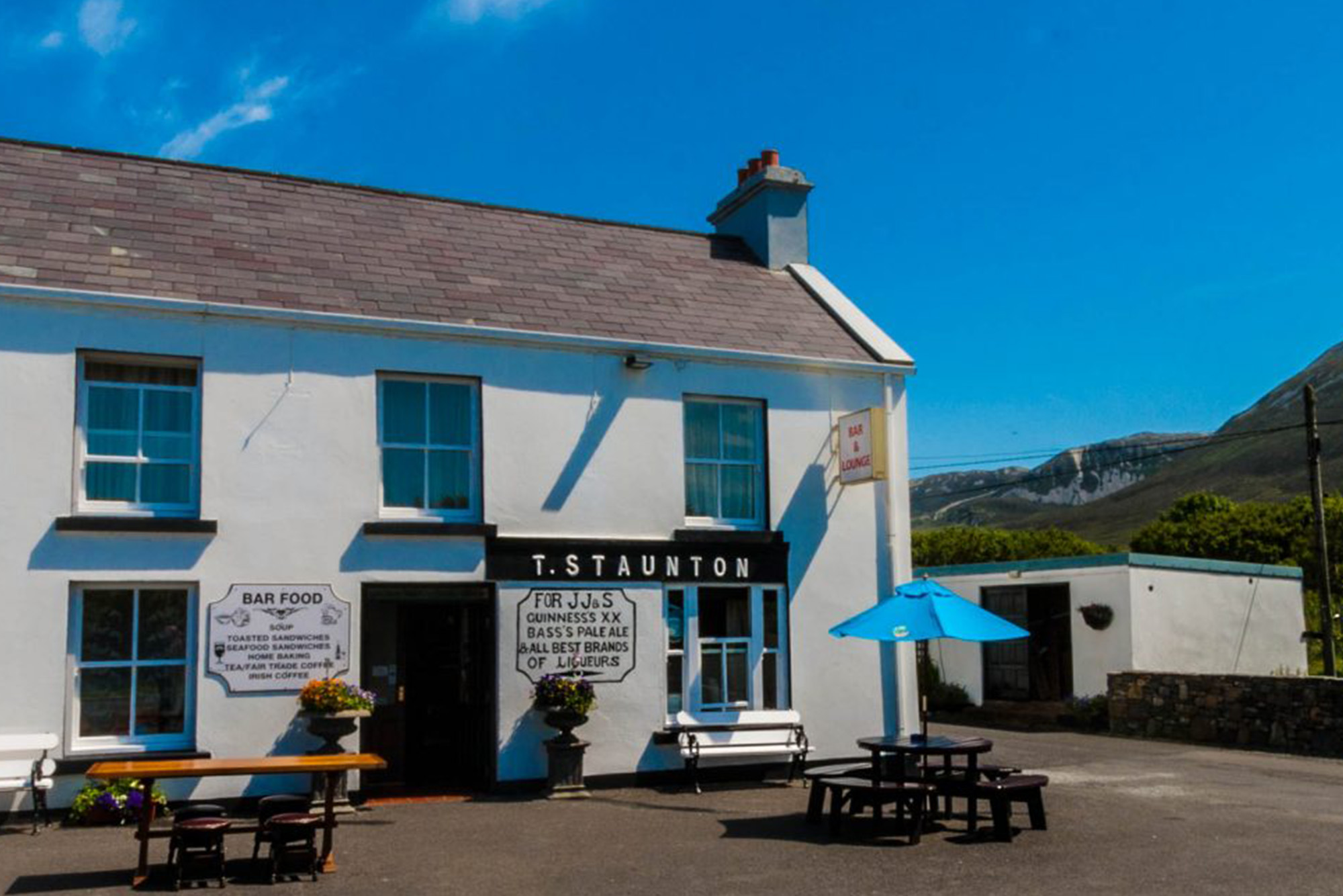 Traditional Irish pub with outdoor seating, scenic mountain views, and blue skies in the background.