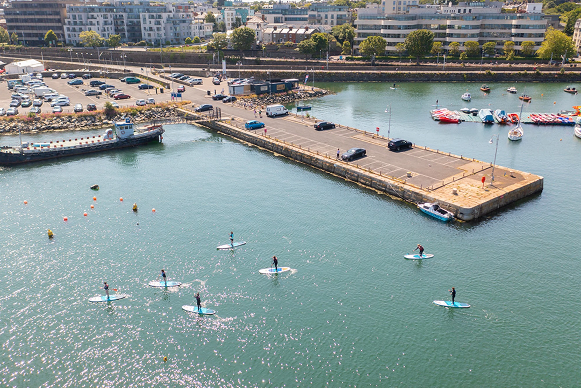 Aerial view of paddleboarders in a coastal marina near a busy car park and docked boats.