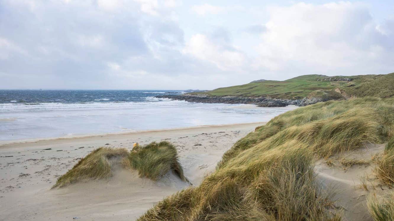 Sandy beach with grassy dunes and rocky coastline under a cloudy sky in Ireland.