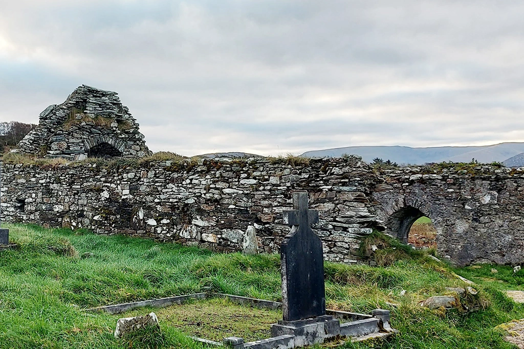 Ancient stone ruins and graveyard on a grassy hillside, with distant mountains under a cloudy sky.