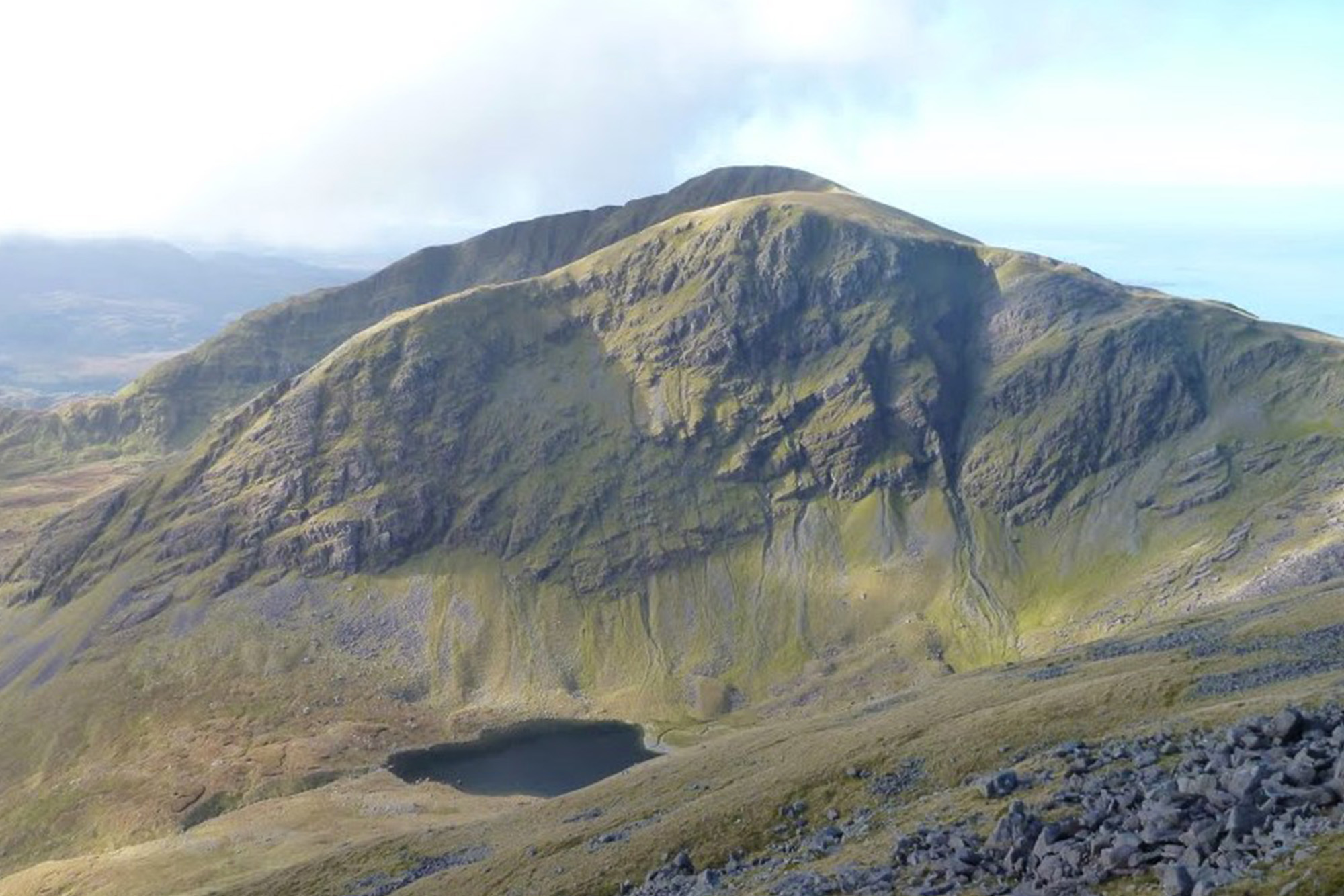 Mountain landscape with green slopes and small lake in Snowdonia, Wales under a cloudy sky.