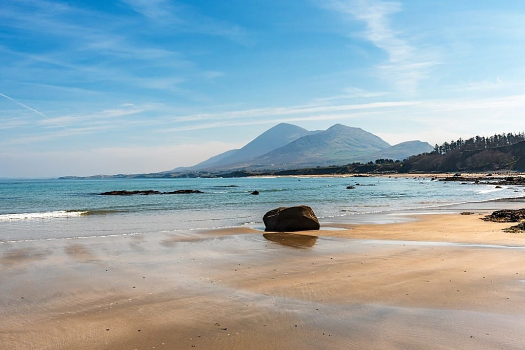 Sunny Irish beach with distant mountains, calm sea, and blue sky; scenic coastal landscape in County Mayo.