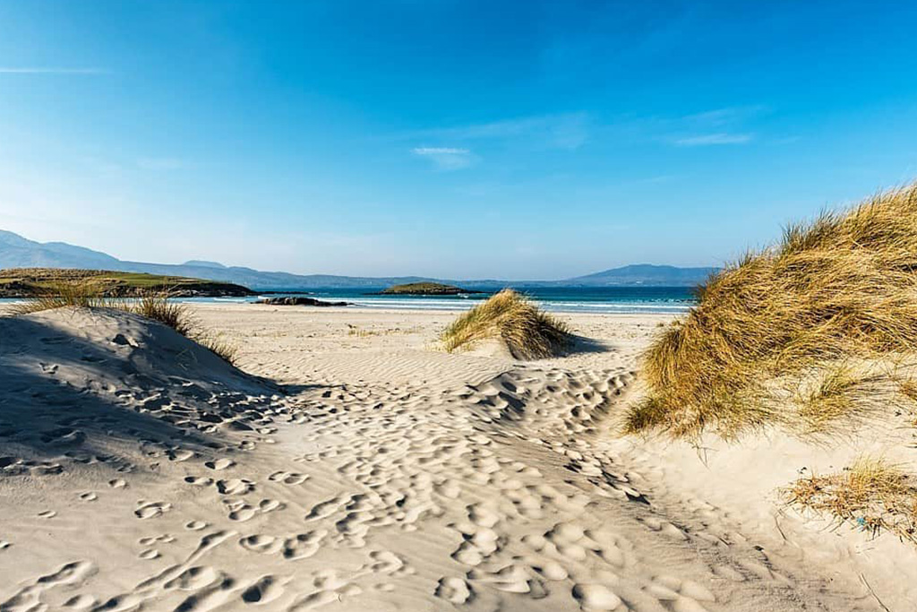 Sunny Irish beach with sand dunes, grassy hills, azure sea and distant mountains under a clear blue sky.