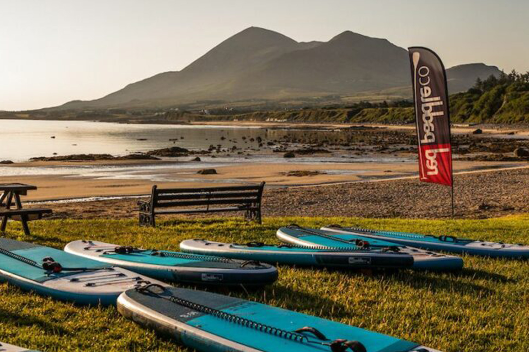 Paddleboards on grassy beach with mountain views, Co. Down, Northern Ireland, under a clear sky.