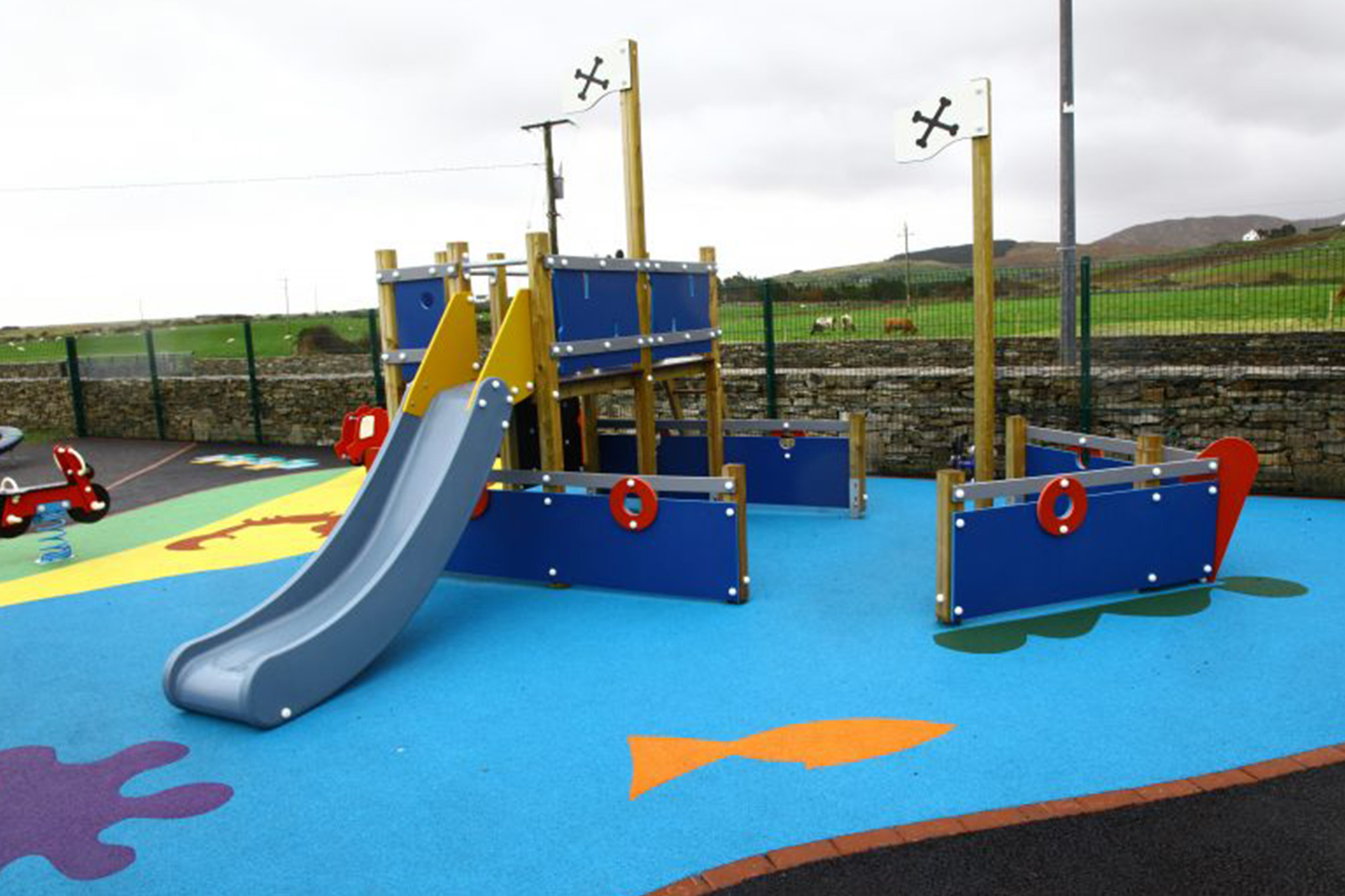 Children's playground with blue pirate ship structure and grey slide on colourful soft surface.